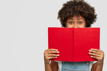 Indoor shot of of cheerful woman covers face with red textbook, has joyful expression, being in high spirit as reads her favourite story, likes fiction, spends leisure with hobby, isolated on white