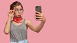 © WHstudio Leushin N - Upset gloomy girlfriend purses lips, makes selfie portrait with modern cell phone, keeps hand on rim of spectacles, models against pink background with blank space for your promotional content