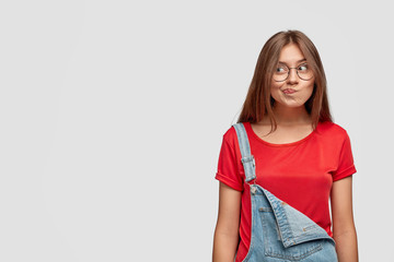 Wall Mural - Horizontal shot of clueless beautiful girl purses lips, has doubtful hesitant facial expression, thinks what to buy, dressed in fashionable denim dungarees, over white background with copy space