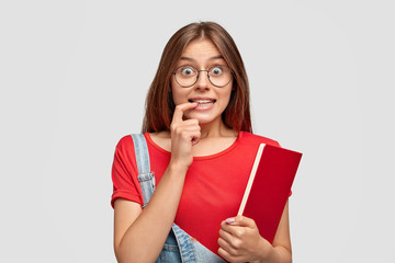 Wall Mural - Pretty student looks with curious expression, eyes full of interest, surprisement, bites fore finger, carries book, listens lecture with curiosity, finds out something new, dressed in stylish outfit