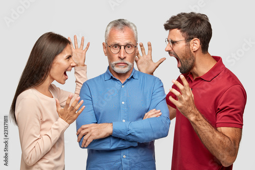 Guilty grey haired bearded senior man keeps arms folded, hears approaches from young colleagues who shout angrily at him. Annoyed woman and man yell at mature father, sort out family relationships