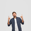 © WHstudio Leushin N - Vertical shot of surprised unshaven guy points with both index fingers upwards, shows free space for your advertising content, keeps mouth opened from amazement. Just look upstairs, its awesome!