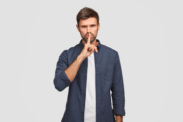 Wall Mural - Photo of serious male keeps index finger on lips as demonstrates hush gesture, asks be quiet, dressed in fashionable clothes, angry as someone tells his secret, isolated over white background