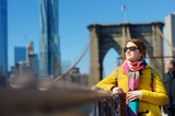 Fototapeta  - Happy young woman tourist sightseeing at Brooklyn Bridge, New York City, at sunny spring day. Female traveler enjoying view of downtown Manhattan.