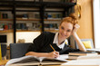 © Drobot Dean - Happy redhead lady student sitting at the table with books in library writing notes.