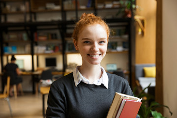 Poster - Happy redhead lady student posing indoors in library holding books.