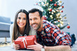 © baranq - Photo of smiling couple looking at camera while relaxing in decorated room exchanging christmas present