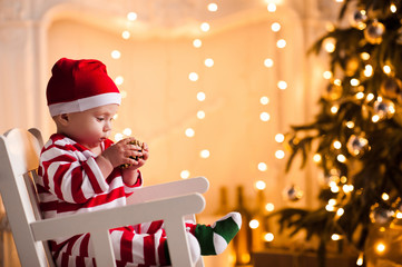 Baby 1 year old wearing santa claus suit sitting in rocking chair over Christmas tree and lights on background in room. Holiday season.