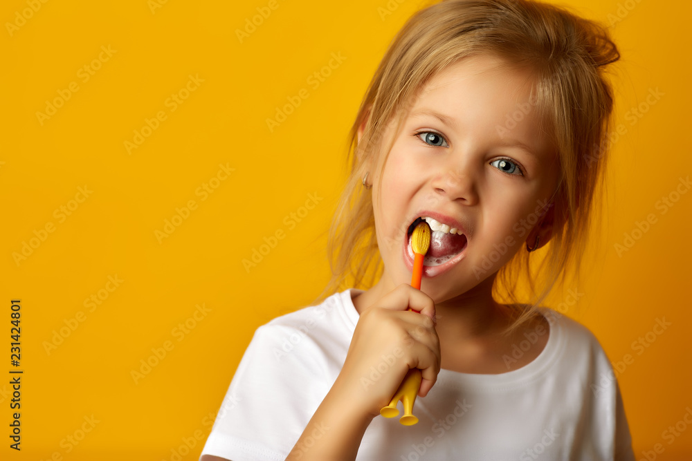 Charming little girl in white t-shirt cleaning teeth with colorful kids toothbrush looking at camera - obrazy, fototapety, plakaty 