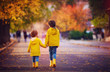 two happy kids, brothers walking together on autumn street in yellow raincoats and rubber boots