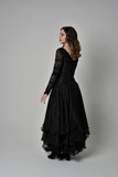 Fototapeta  - full length portrait of brunette girl wearing long black lace gown with corset.  standing pose with back to the camera, grey studio background.