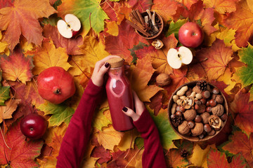 Wall Mural - Autumn vegan and vegetarian food concept - apples, pomegranate, nuts, spices. Picnic time. Female hands holding bottle of red summer drink - smoothie or juice over colorful leaves background. Top view