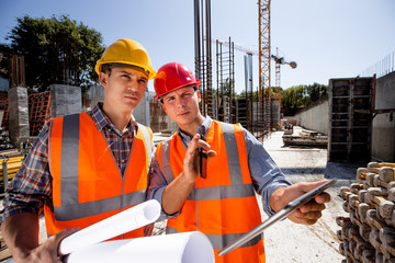 Architect  and structural engineer dressed in orange work vests and  helmets discuss a building project on the tablet on the open air building site with construction material