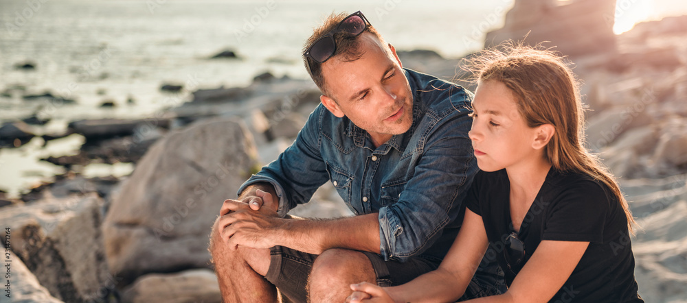 Father and daughter sitting on a rocky beach and talking - obrazy, fototapety, plakaty 