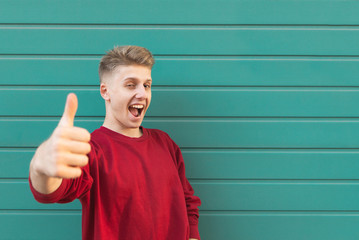 Expressive young man stands on the background of a turquoise wall, showing a thumbs up and looking into the camera. Emotional happy man shows likes.