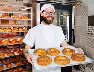 attractive Baker in white uniform holding a tray with freshly baked bagels with sesame and poppy seeds on the background of a bread factory or bakery