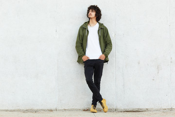 Full length shot of pensive teenger dressed in fashionable anorak, keeps hands in pockets, focused aside, contemplates about something, isolated over white concrete wall. Youth, lifestyle concept