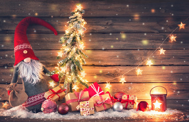 Wall Mural - santa claus or dwarf holds a fir tree with christmas lights surrounded by gift boxes and glowing lan