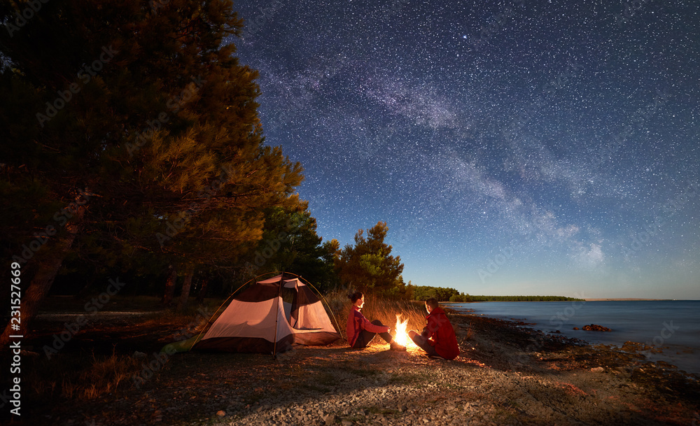 Night camping on shore. Man and woman hikers having a rest in front of tent at campfire under evening sky full of stars and Milky way on blue water and forest background. Outdoor lifestyle concept - obrazy, fototapety, plakaty 