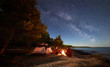 Leinwandbild Motiv Night camping on shore. Man and woman hikers having a rest in front of tent at campfire under evening sky full of stars and Milky way on blue water and forest background. Outdoor lifestyle concept