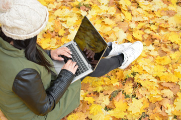 Wall Mural - Girl in hipster with laptop in autumn park. A woman in a cap using a laptop while sitting on fallen leaves. Freelancer in the hat uses remote communication technology. Remote work. View from above