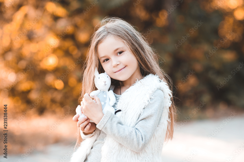 Pretty child girl 5-6 year old holding cute toy rabbit outdoors. Looking at camera. Childhood. Stock Photo | Adobe Stock