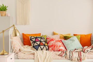 Patterned and colorful pillows on comfortable white bed in stylish bedroom interior with golden lamp on bedside table and handmade macrame on white wall, real photo with copy space