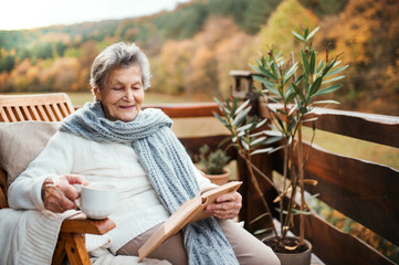 An elderly woman reading book outdoors on a terrace on a sunny day in autumn.