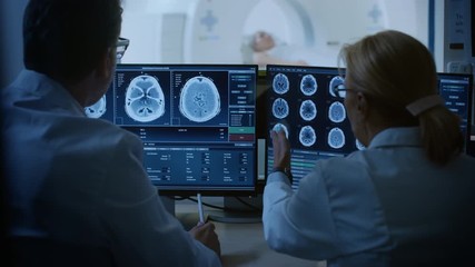 Wall Mural - In Control Room Doctor and Radiologist Discuss Diagnosis while Watching Procedure and Monitors Showing Brain Scans Results, In the Background Patient Undergoes MRI or CT Scan Procedure.