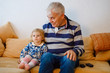 © Irina Schmidt - cute little toddler girl and grandfather watching together tv show. Baby granddaughter and happy retired senior man sitting together at home with cartoons on television.