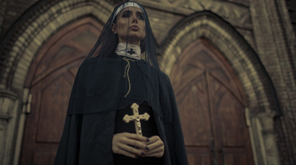 Young nun is standing and praying with cross in her hands on temple background.