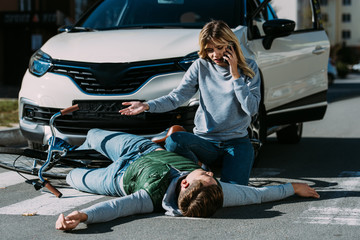 Wall Mural - woman calling emergency and looking at injured cyclist lying on road after traffic collision