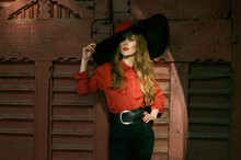 Attractive Slim Woman In A Wide-brimmed Hat, A Red Blouse And Black Pants Posing Against A Vintage Red Door. Fashionable Photo.
