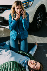 Wall Mural - high angle view of scared young woman calling emergency while injured man lying on road after traffic accident