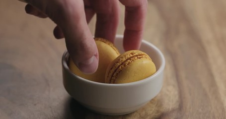 Wall Mural - Slow motion man hand put orange macarons in white bowl on wood table