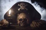 Human skull with pirate captain hat above, burning candle, seashell and mooring rope on the wooden table in the mystic smoke.