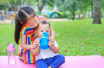 Adorable Asian sister sitting on pink mattress mat take care her little brother to drinking water from Baby sippy cup with straw in the park garden.