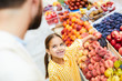 © pressmaster - Happy excited beautiful daughter in yellow dress standing at food stall and pointing at peaches while asking father to buy it, she wants fruits