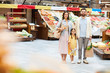 © pressmaster - Positive beautiful young family in casual clothing standing in farmers market and holding shopping bags while looking at camera