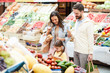 © pressmaster - Serious concentrated little daughter with pony tails standing at food shelves and scanning price tag using tablet in food store, cheerful parents looking at tablet screen while making purchases