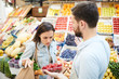 © pressmaster - Puzzled frowning young female customer with shopping bag on shoulder displeased with quality of products and looking at strawberry shown by grocer in farmers market