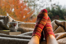 Woman's Feet Wearing Handmade Knitted Colorful Wool Socks Next To A Hot Cup Of Tea In A Cozy Decor. Keeping Warm On A Cold Day Concept. 