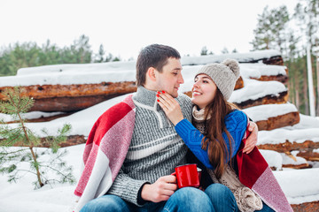 Wall Mural - Young loving couple dressed in blue sweater sitting with red coffee cups on the firewood in winter forest