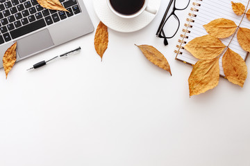 White desk copy space seen from above with a laptop keyboard, notebook, glasses, coffee, pen and yellow autumn leaves