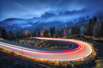 Wall Mural - Blurred car headlights on winding road in mountains with low clouds at night in autumn. Spectacular landscape with asphalt road, light trails, foggy forest, rocks and blue sky. Car driving on roadway
