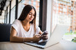 © dianagrytsku - Young beauty woman sitting with phone in hands typing in front of laptop in modern cafe