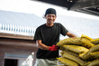 © Mint Images - Japanese farmer wearing black cap standing next to stack of yellow plastic sacks, smiling at camera.