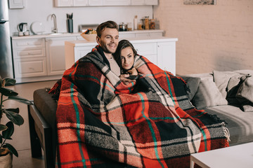 happy young couple relaxing on couch and watching tv together while covering with plaid