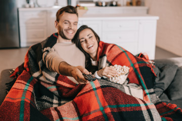 happy young couple watching tv with popcorn on couch and covering with plaid