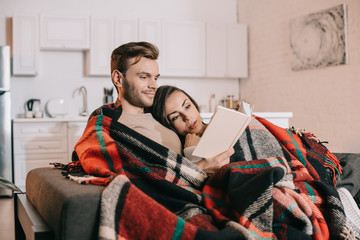 Wall Mural - happy young couple reading book together on couch under plaid at home